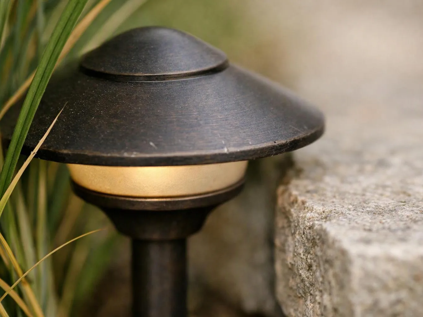 A black garden path light glows beside a stone edge and green leaves outdoors
